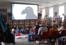 Kinder vor einemgroßen Fernseher auf dem ein Zeichentrick-Rabe zu sehen ist.